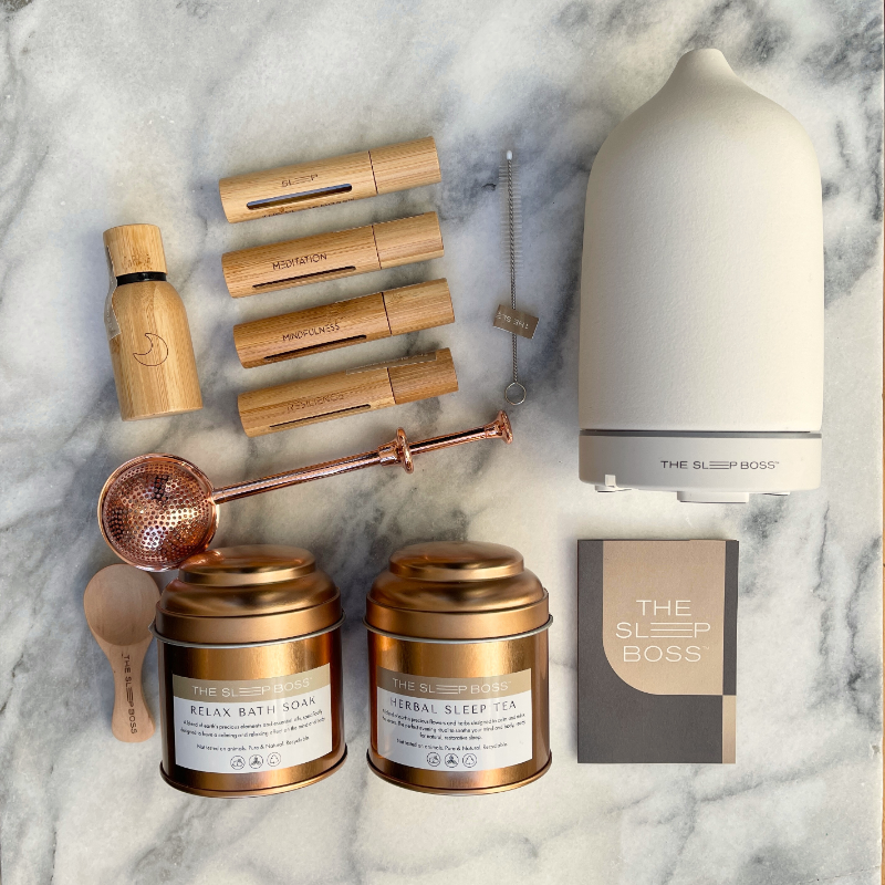 A collection of Herbal Sleep Tea, Essential Oil Rollers, a white ceramic diffuser and Sleep pure oil in a gift set on marble table.
